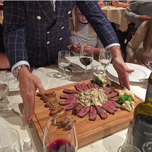 A person presents a wooden platter of sliced steak, garnished with vegetables and sauce, at a dining table.