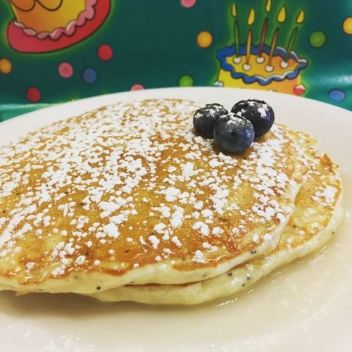Two fluffy pancakes dusted with powdered sugar, topped with three blueberries, against a colorful birthday-themed background.