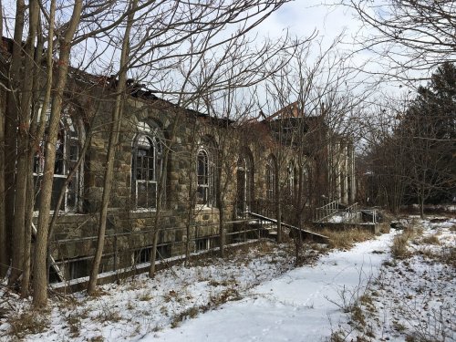 Abandoned stone building surrounded by overgrown trees and snow, with broken windows and a deteriorating roof.