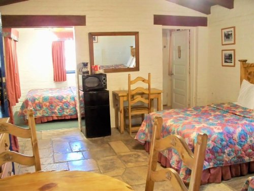Cozy hotel room with two beds, a small dining table, and a kitchenette area, featuring floral bedspreads and warm lighting.