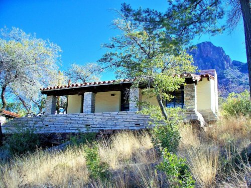 A rustic house with stone walls, surrounded by trees and grass, set against a backdrop of mountains and clear blue sky.