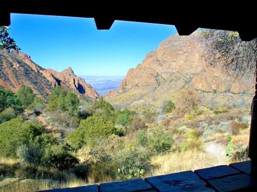 View of a mountainous landscape with greenery, framed by a rustic shelter, under a clear blue sky.