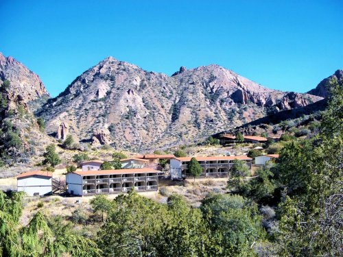 A scenic view of mountains with a cluster of buildings nestled among trees in a sunny landscape.