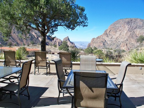 A patio with empty tables and chairs, overlooking a scenic view of mountains and clear blue sky.