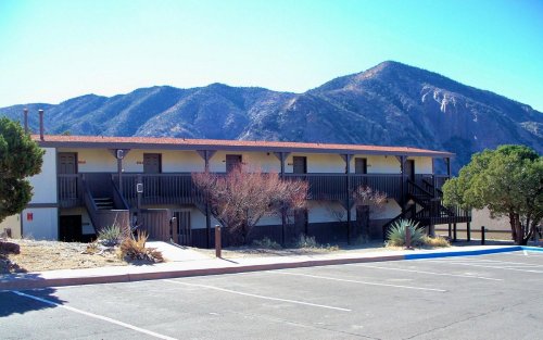A two-story building with a balcony, surrounded by desert vegetation and mountains in the background under a clear blue sky.