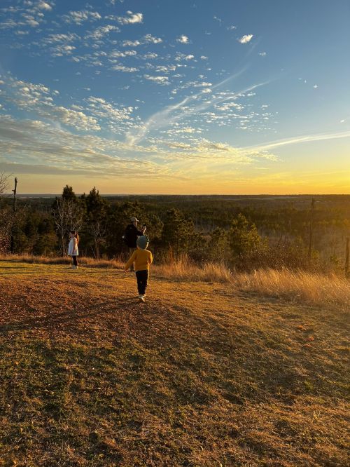 Two people walk on a grassy hill at sunset, with a vast landscape and colorful sky in the background.