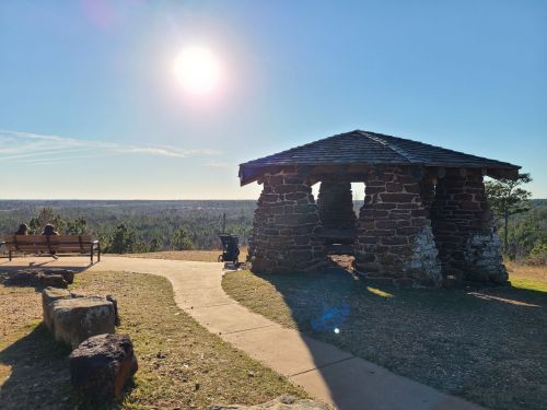 A stone gazebo overlooks a scenic landscape under a bright sun, with a path and benches nearby.