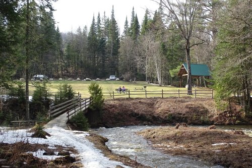 A serene landscape featuring a stream, a wooden bridge, and a picnic area surrounded by trees and a grassy field.