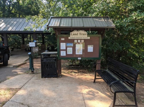 Information board for Land Trust at a nature preserve, with a bench and trash can nearby, surrounded by greenery.