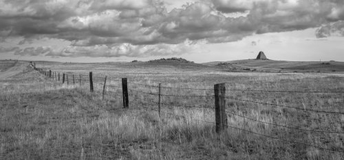 A black and white landscape featuring a barbed wire fence, open fields, and a distant hill under a cloudy sky.