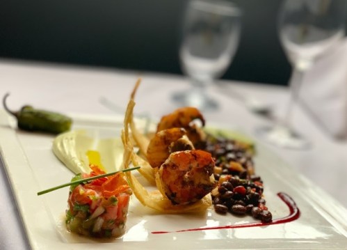 A gourmet plate featuring grilled shrimp, black beans, and a fresh salsa garnish, elegantly presented on a white dish.