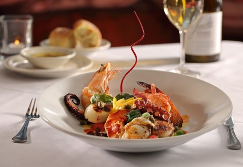 A gourmet seafood dish featuring shrimp, octopus, and vegetables, served with a glass of white wine.