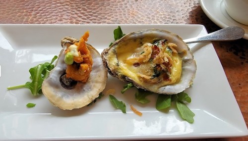 Two oysters on a plate, one raw with toppings and one baked with a golden crust, garnished with arugula.