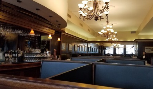 Interior of a restaurant featuring dark wood decor, chandeliers, and a bar with wine glasses and bottles.