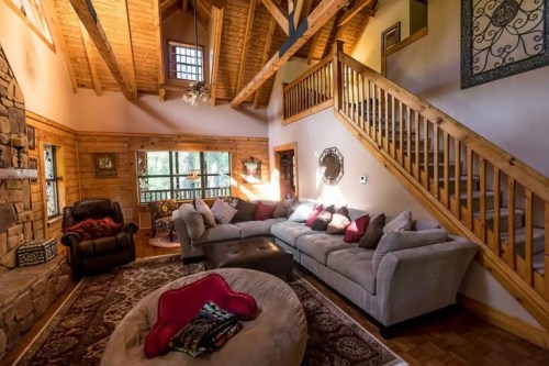 Cozy living room with a stone fireplace, wooden beams, a sectional sofa, and a staircase leading to an upper level.