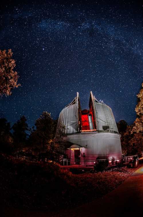 A telescope dome illuminated at night, surrounded by trees under a starry sky.