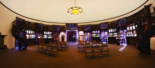 A circular library interior with wooden shelves, displays, and chairs arranged for an event under a dome ceiling.