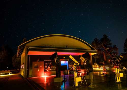 A dark sky filled with stars above the Lowell Observatory, featuring telescopes and illuminated pathways.