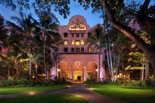 A grand hotel entrance surrounded by lush greenery and palm trees, illuminated at dusk.