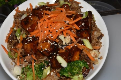 A bowl of fried rice topped with carrots, broccoli, green onions, and glazed tofu, garnished with sesame seeds.