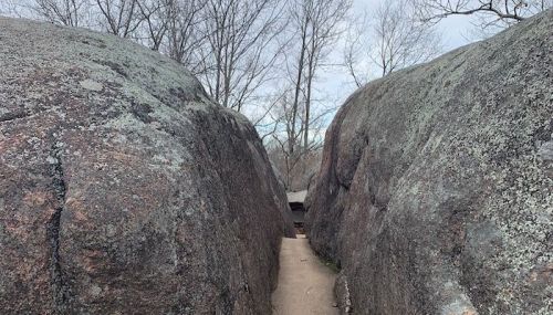 A narrow path between two large, moss-covered boulders, surrounded by bare trees and a cloudy sky.