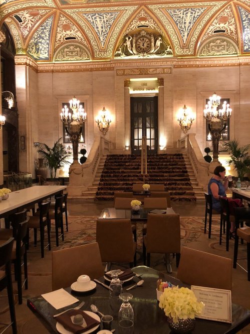 Elegant restaurant interior with ornate ceiling, chandeliers, and a grand staircase, featuring tables and floral arrangements.