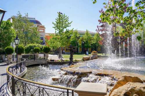 A serene park scene featuring a fountain, lush greenery, and a winding pathway beside a flowing stream.