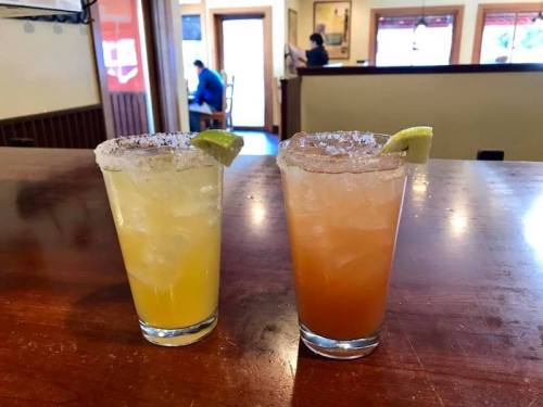 Two cocktails with salted rims and lime wedges, one yellow and one orange, on a wooden table in a restaurant.