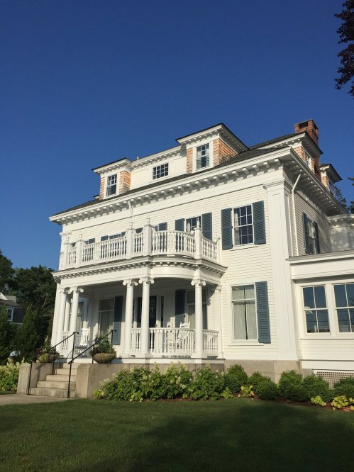 A large, elegant white house with blue shutters and a wraparound porch, set against a clear blue sky.