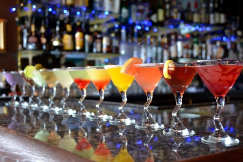 A colorful array of cocktails in elegant glasses lined up on a bar, with a blurred background of bottles.