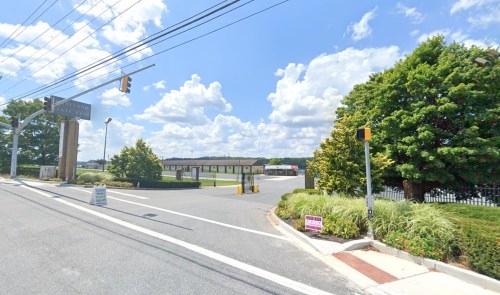 A street view showing a traffic light, green trees, and a clear sky with clouds, leading to a parking lot.