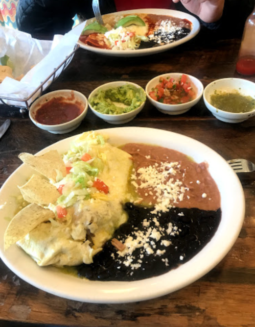 A plate of enchiladas topped with lettuce and tomatoes, served with refried beans, black beans, and various salsas.
