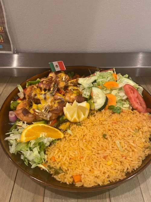 A colorful plate featuring grilled chicken, rice, salad, and garnishes like lemon and orange slices, with a small Mexican flag.