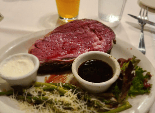 A plate with a large slice of prime rib, sides of asparagus, salad, and dipping sauces, with a drink in the background.