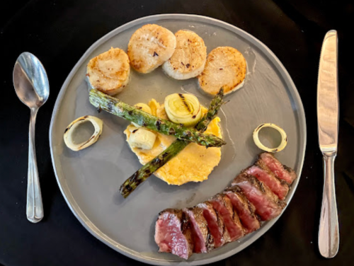 A gourmet plate featuring scallops, asparagus, steak slices, and a creamy base, elegantly arranged on a gray dish.