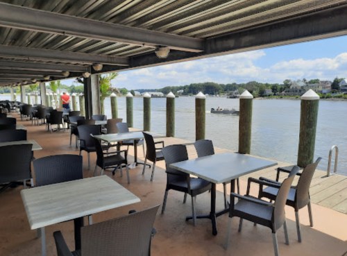 Outdoor dining area by a river, featuring empty tables and chairs under a covered patio with a boat in the distance.