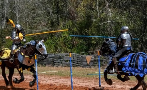 Two armored knights on horseback engage in a jousting match, with lances aimed at each other in a grassy arena.