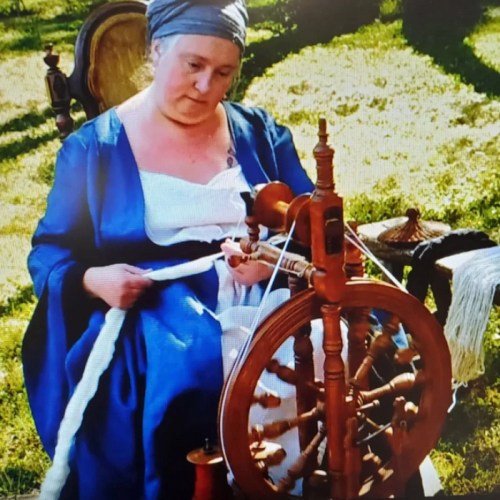 A woman in historical attire spins yarn on a wooden spinning wheel outdoors, surrounded by greenery.