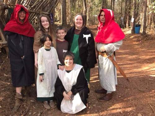 A group of six people in medieval-style costumes posing together in a wooded area.
