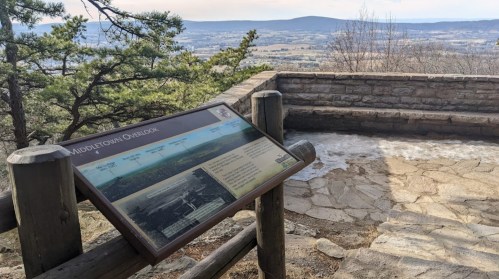 A scenic overlook with a sign detailing the view, surrounded by trees and rocky terrain. Mountains are visible in the distance.
