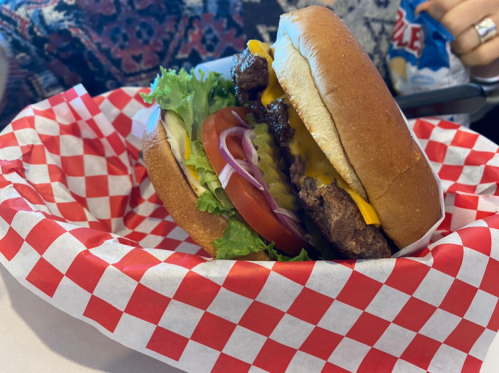 A juicy hamburger with lettuce, tomato, pickles, and cheese in a bun, served in a red and white checkered basket.