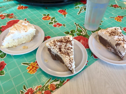 Three slices of pie on white plates: a lemon meringue, a chocolate cream, and a coconut cream, on a floral tablecloth.