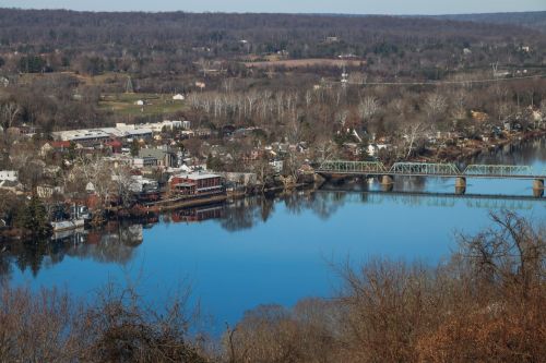 A scenic view of a river town with a bridge, surrounded by trees and hills, reflecting in the calm water below.