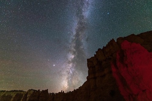 A starry night sky with the Milky Way above rugged rock formations, illuminated by a soft red glow.