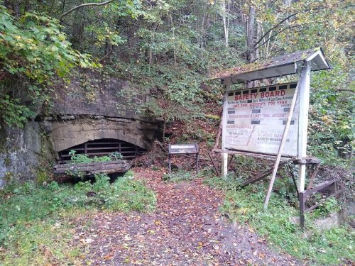 An old stone entrance surrounded by trees, with a safety board sign and a weathered cart nearby.
