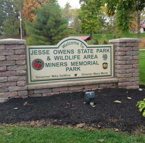 Sign at Jesse Owens State Park and Wildlife Area, featuring park name and officials' names, surrounded by greenery.