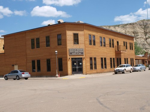A two-story wooden building with large windows, parked cars, and a blue sky with clouds in the background.