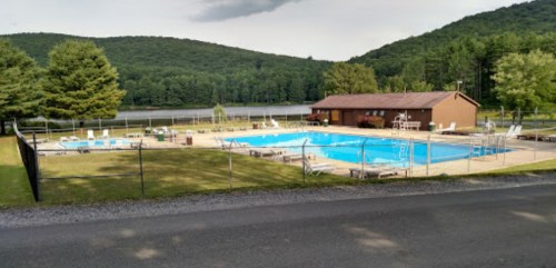 A fenced swimming pool surrounded by lounge chairs, with a cabin and mountains in the background.