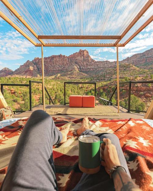 A person relaxes on a bed with a blanket, holding a drink, overlooking a scenic mountain view.