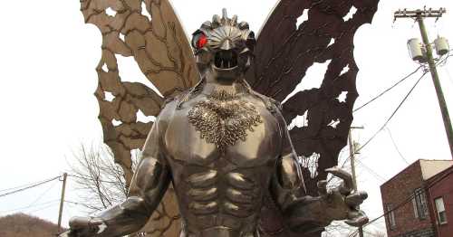 A large metallic statue of a creature with wings, red eyes, and a muscular build, set against a cloudy sky.